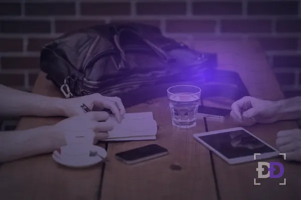 Two people sit at a wooden table, one taking notes in a notebook while the other has a phone and a tablet in front of them.