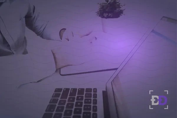 A man writes notes in a notebook next to an open laptop showing a document on a white desk.